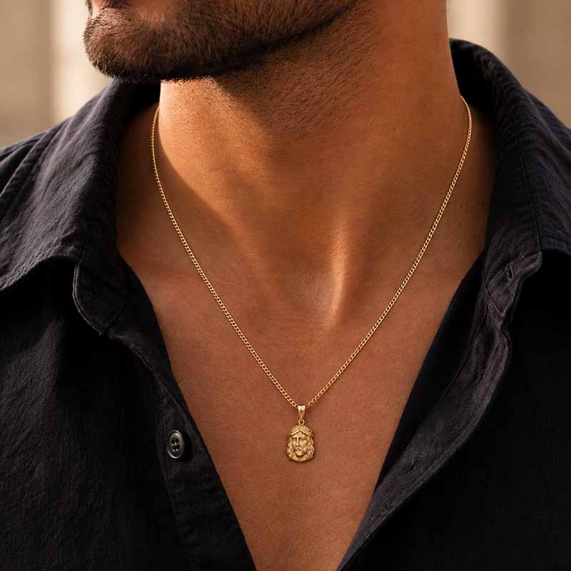 A close-up of a man wearing a delicate gold chain necklace with a small, detailed gold pendant, styled with a black button-down shirt.