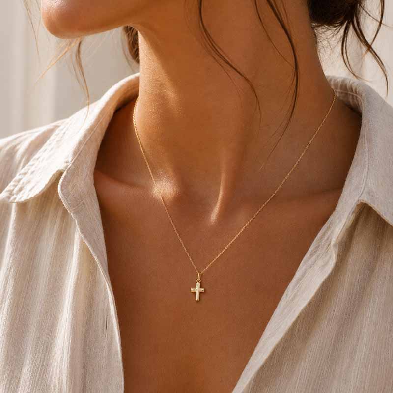 A close-up lifestyle shot of a model wearing a delicate gold cross pendant necklace on a fine chain, paired with a light beige linen shirt.