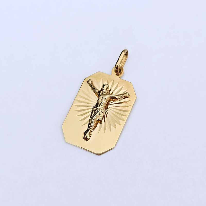 A gold crucifix pendant charm featuring an engraved figure of Jesus Christ on a sunburst background, shown on a light lavender background.