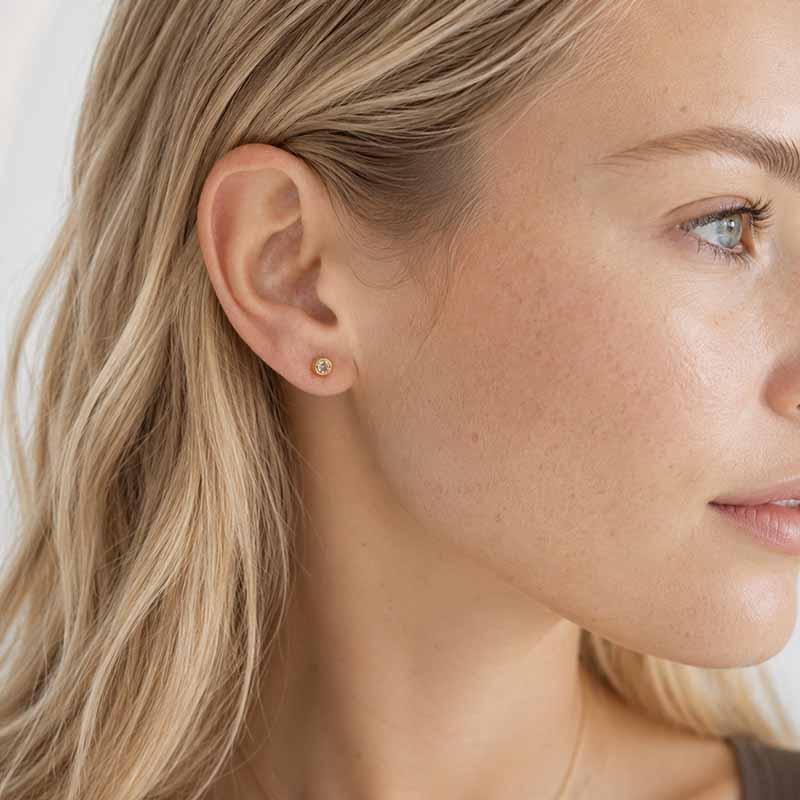 A close-up side profile of a woman with blonde hair wearing a small gold stud earring with a clear gemstone, set against a soft light gray background.