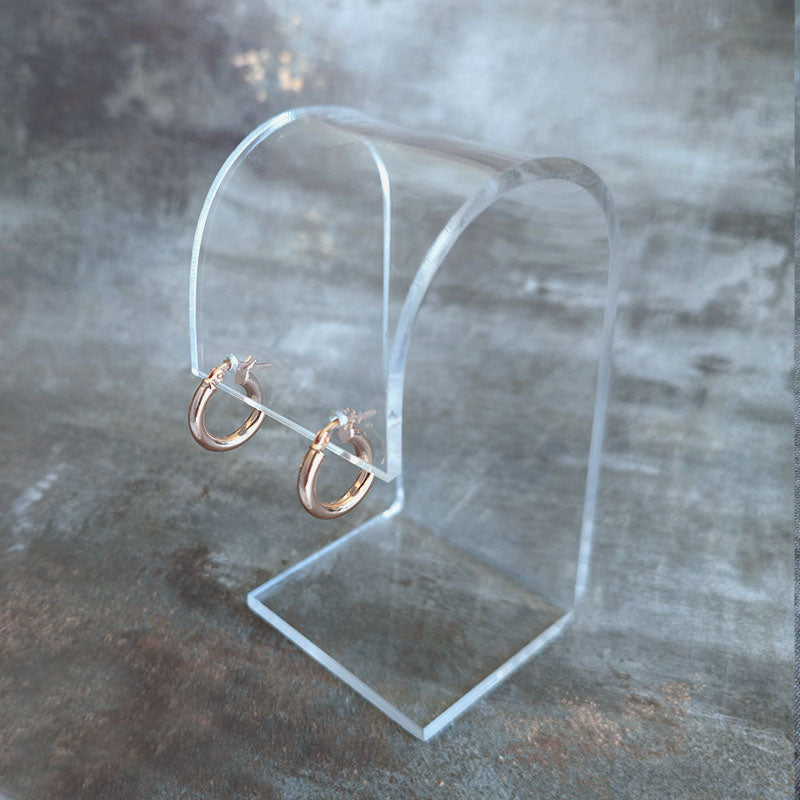 Rose gold hoop earrings on a clear acrylic stand against a textured gray background