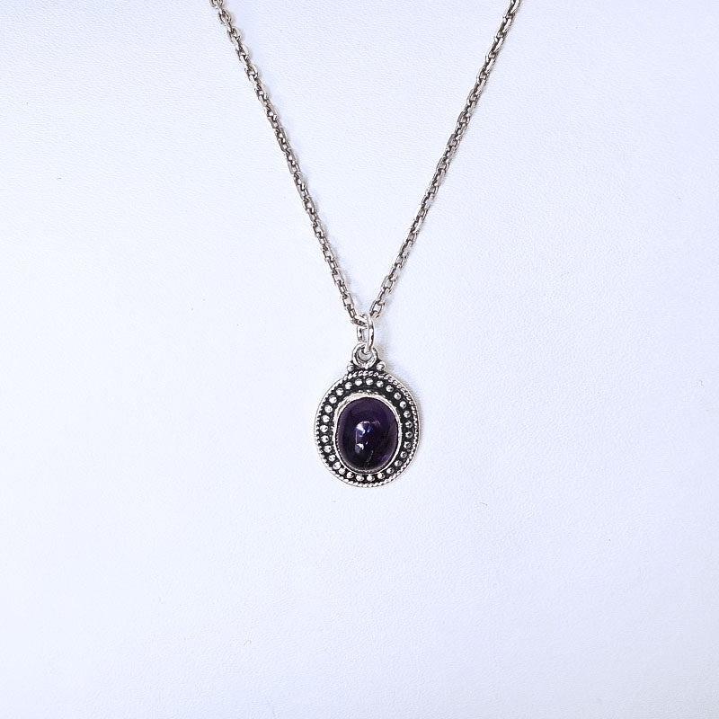 A silver-tone chain necklace with an oval pendant featuring a dark purple cabochon gemstone surrounded by a silver beaded halo, displayed against a plain white background.
