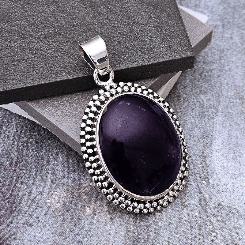 A silver oval pendant featuring a dark purple amethyst cabochon stone, surrounded by a textured beaded silver border, displayed on a gray textured surface with a dark gray presentation box.