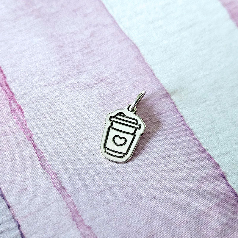 Silver pendant shaped like a coffee cup with a heart on a textured pink and white background