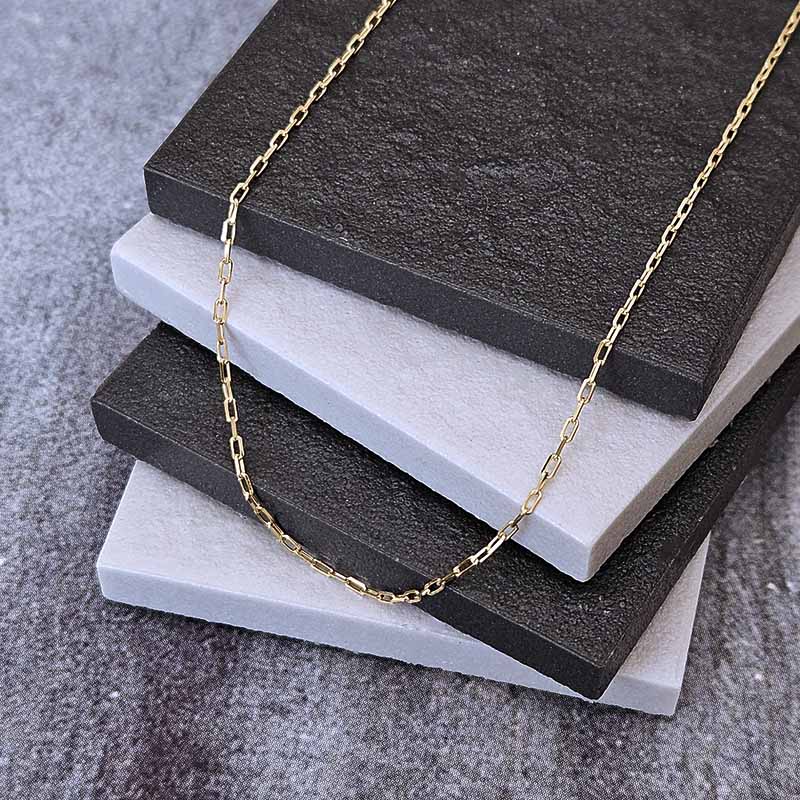 A delicate gold chain necklace draped over a stack of textured square tiles in dark charcoal and light gray, set on a gray fabric surface.