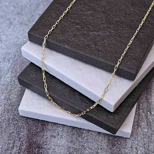 A delicate gold chain necklace draped over a stack of textured square tiles in dark charcoal and light gray, set on a gray fabric surface.