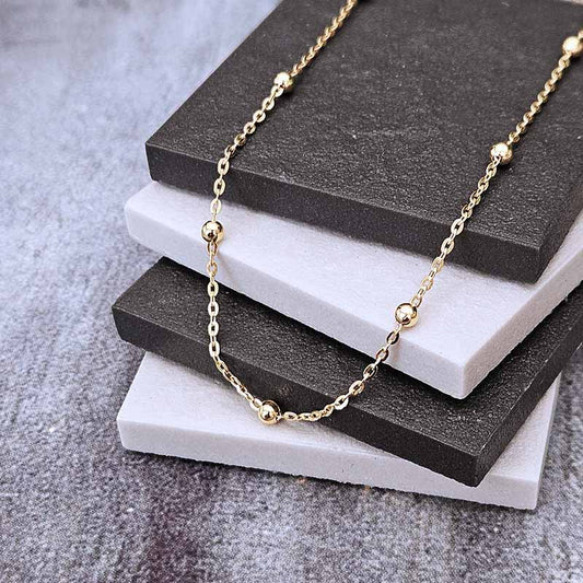 A delicate gold chain necklace with small spherical bead accents, draped over a stack of dark gray and white display blocks on a textured gray surface.