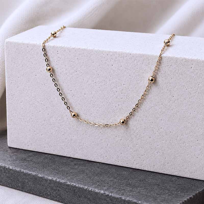 A delicate gold chain necklace with small spaced beads draped over a textured white rectangular block, set against a soft white fabric background.