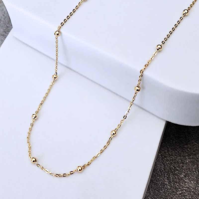 A delicate gold chain necklace with small, evenly spaced polished bead accents, draped over a white jewelry display stand.