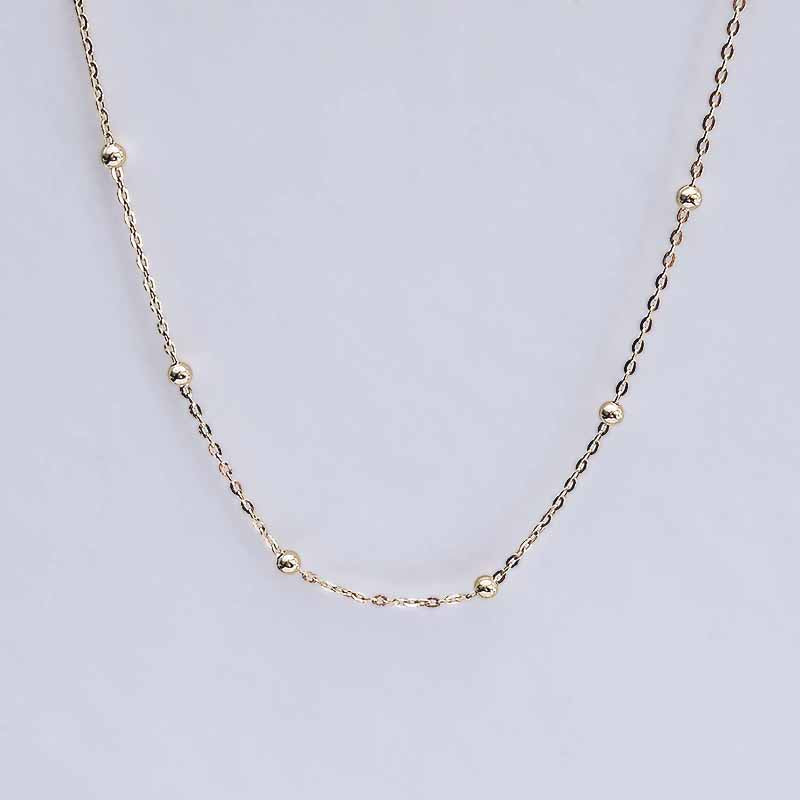 A delicate gold chain necklace featuring small, evenly spaced polished bead accents, displayed against a light gray background.