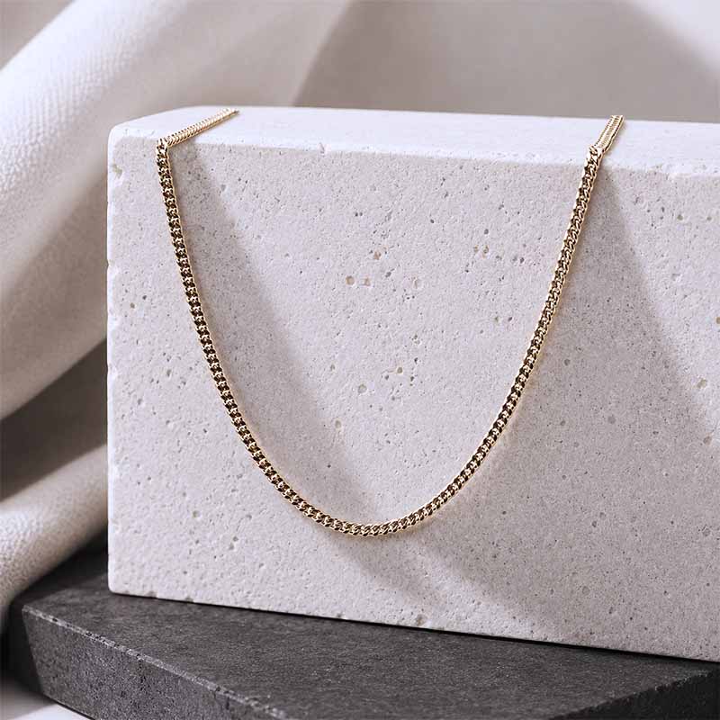 A delicate gold chain necklace draped over a textured white stone block, set against a soft beige fabric backdrop and a dark gray slate base.
