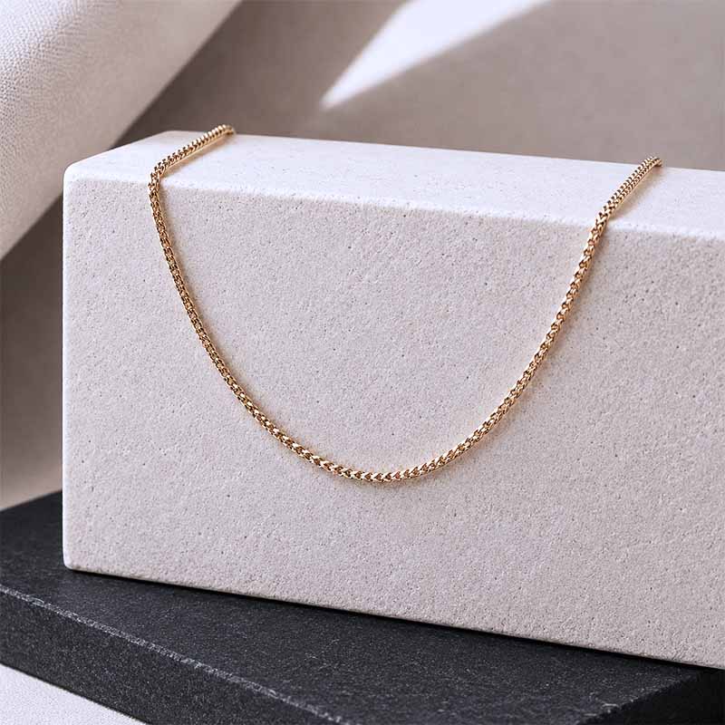 A delicate gold chain necklace draped over a textured white display block, set against a neutral beige background with a dark gray surface below.