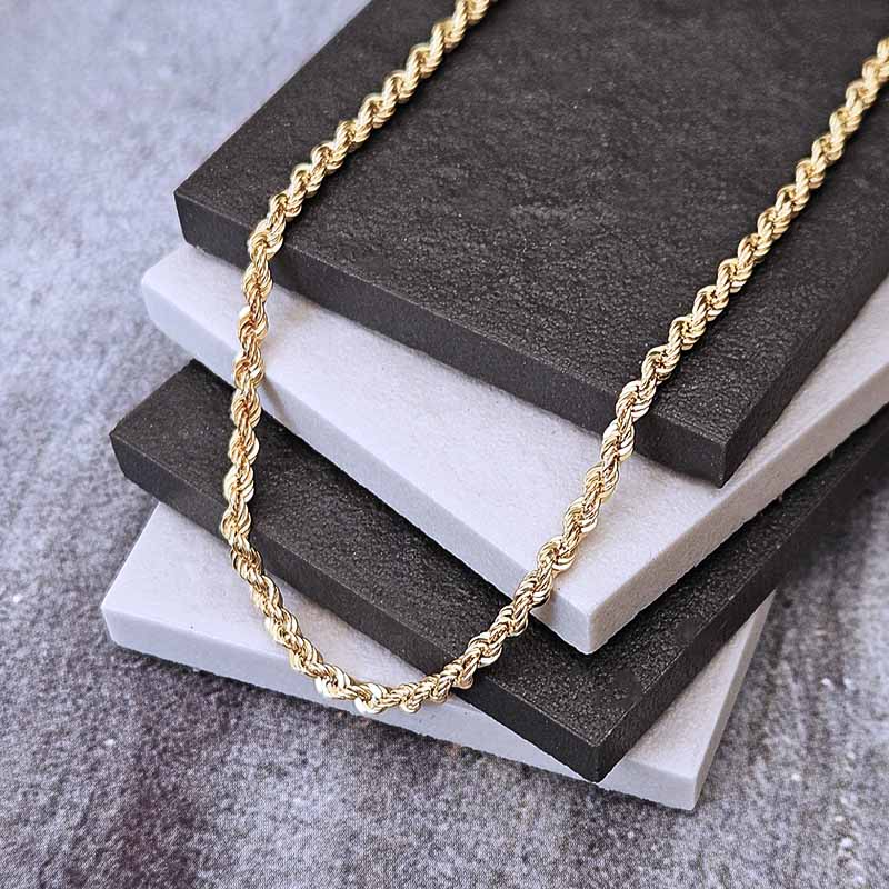 A gold twisted rope chain necklace draped over a stack of dark and light gray textured display blocks on a gray fabric surface.