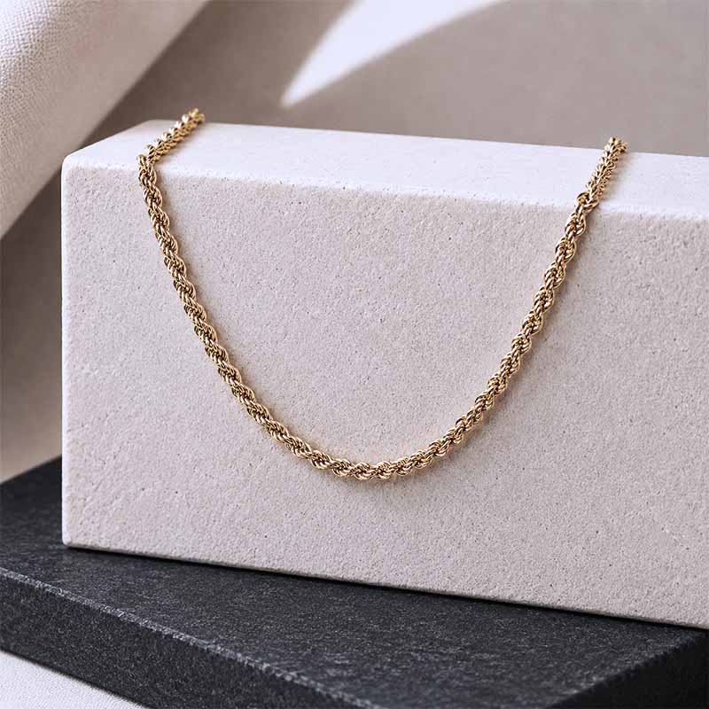 A gold rope chain necklace draped over a textured white display block, set against a neutral beige background with a dark gray surface below.