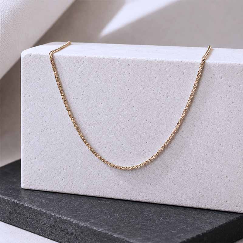 A delicate gold chain necklace draped over a textured white rectangular block, set against a neutral beige and dark gray background.