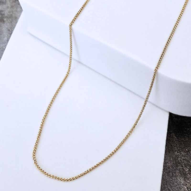A delicate gold chain necklace draped over a white display block, photographed against a clean white background with a small dark textured area at the lower right.