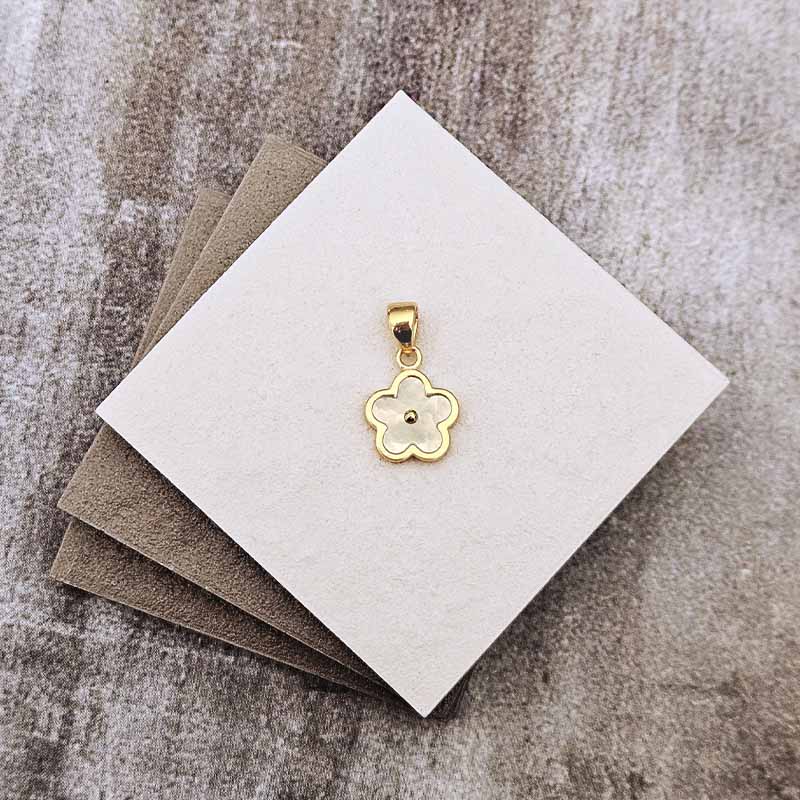 A small gold flower-shaped pendant with a central bead, displayed on a white square card resting on brown textured paper over a gray stone-like surface.