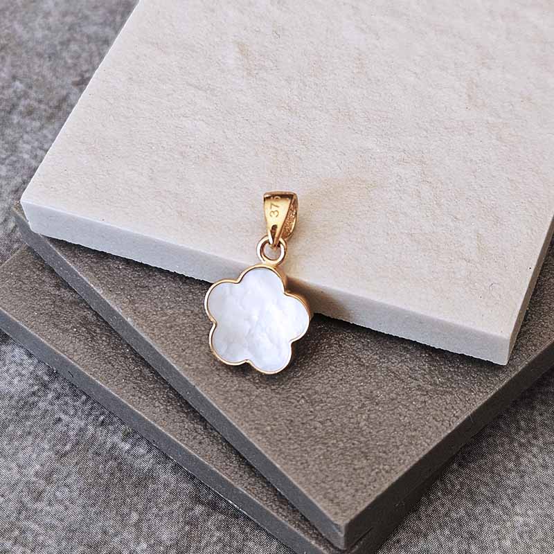 A gold four-petal flower pendant with a white mother-of-pearl inlay, resting on stacked stone tiles.