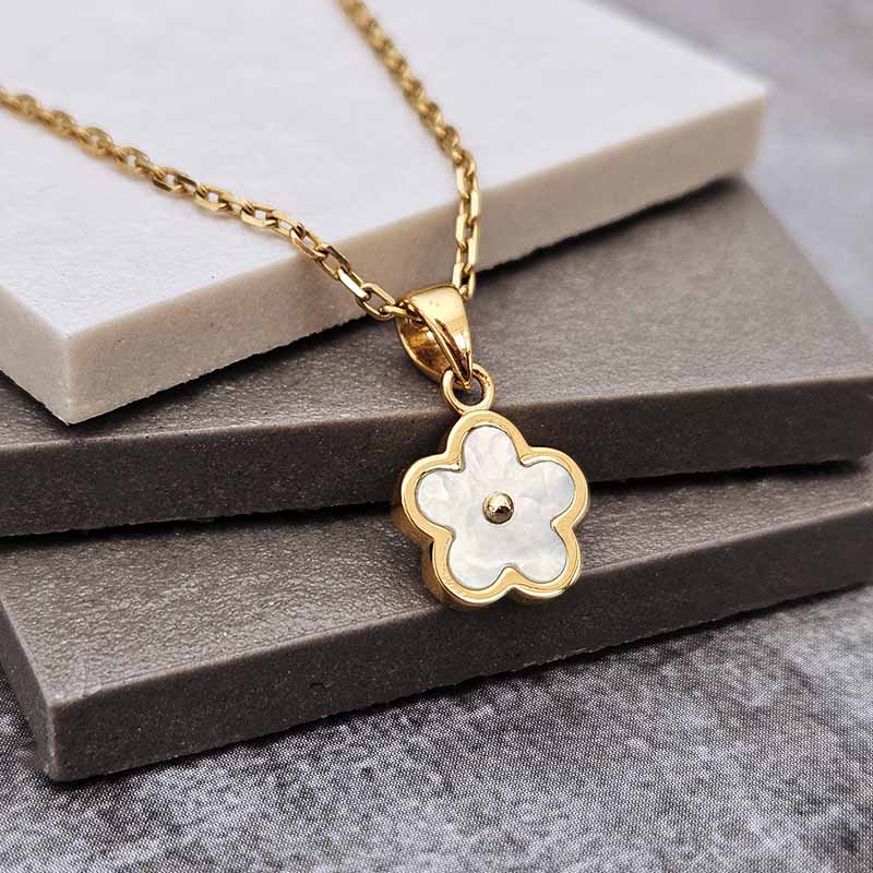 A gold-tone chain necklace with a five-petal flower pendant featuring a white inlay and a small central gold dot, displayed on stacked gray and white stone blocks.