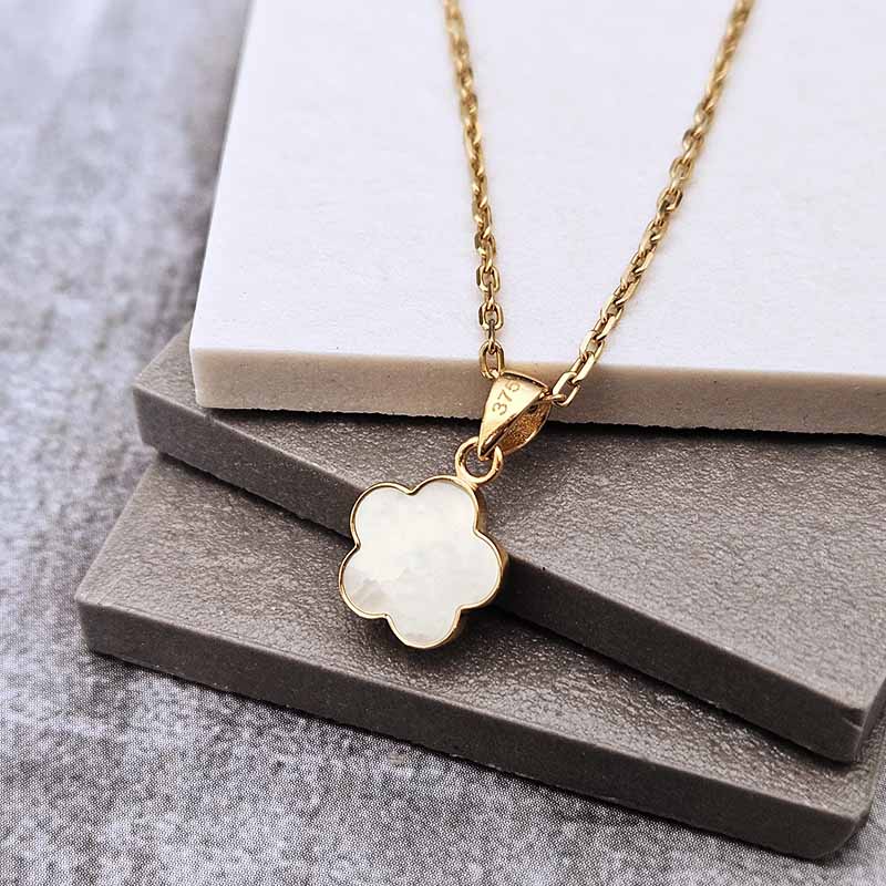 A gold chain necklace with a clover-shaped pendant featuring an iridescent white center, displayed on stacked gray and white stone blocks.