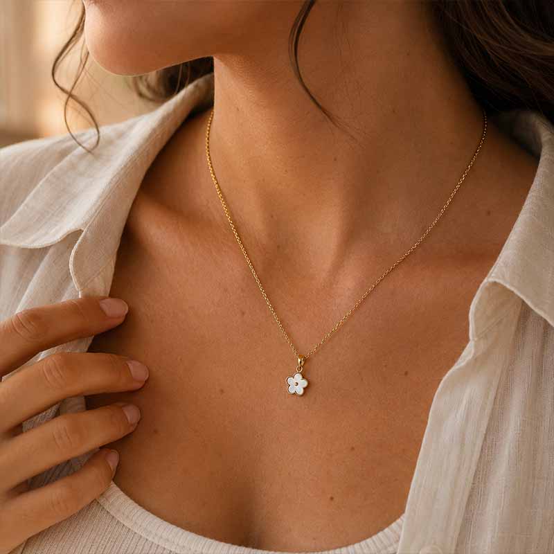 A close-up lifestyle shot of a woman wearing a delicate gold chain necklace with a small white flower pendant, styled with a white linen shirt.