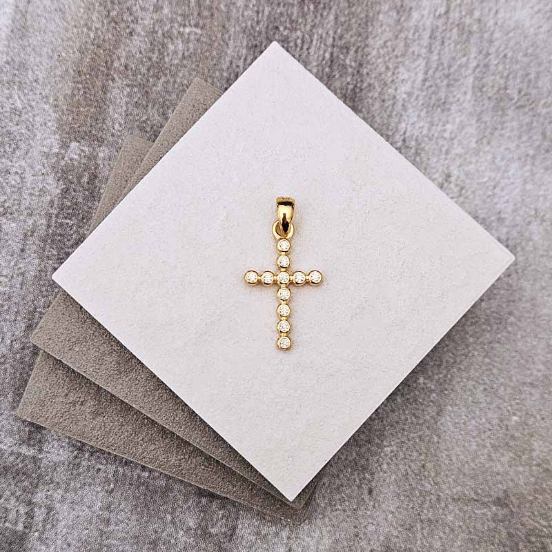 A small gold cross pendant with clear stones, displayed on a white square card resting on gray textured paper.