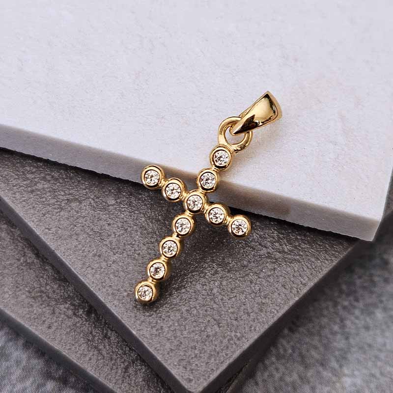 A gold cross pendant adorned with small clear stones, resting on a textured gray surface with a white block in the background.