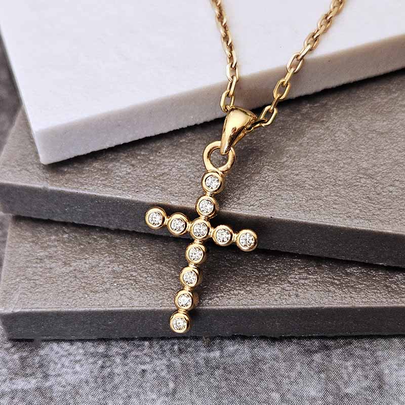 A gold cross pendant necklace adorned with small clear stones, resting on stacked gray stone slabs with a white marble block in the background.