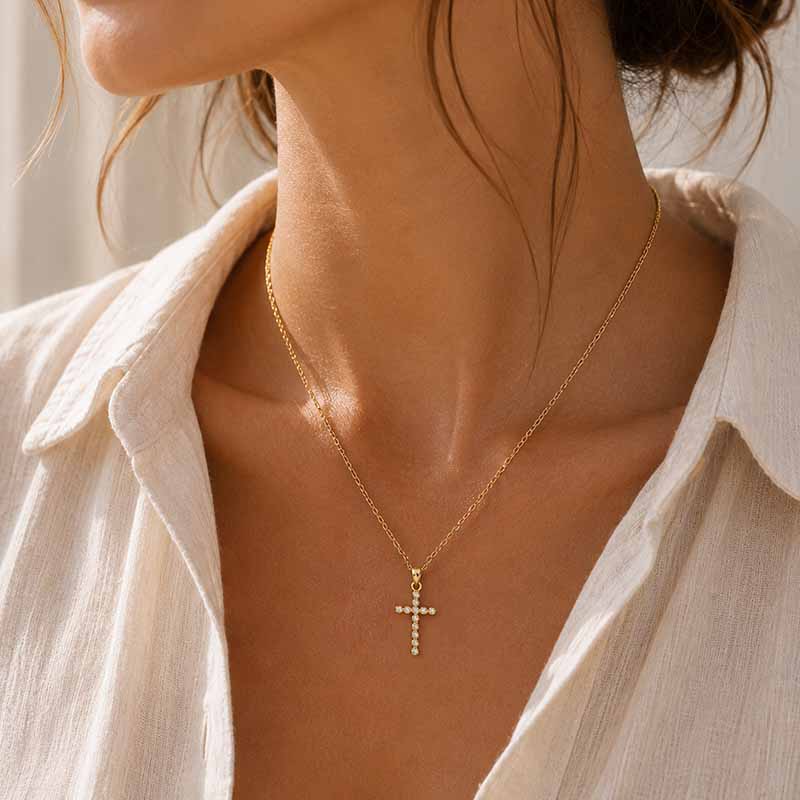 A close-up of a woman's neck and upper chest wearing a delicate gold cross pendant necklace with small clear stones, paired with an off-white collared shirt.