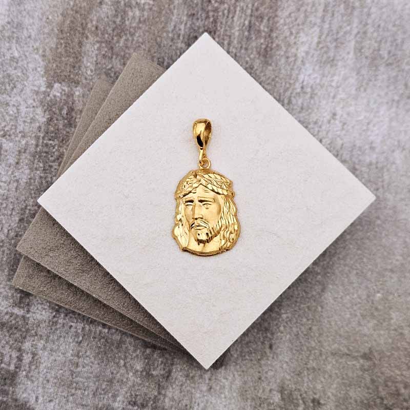A gold Jesus face pendant with a crown of thorns, displayed on a white square card resting on a gray textured surface.