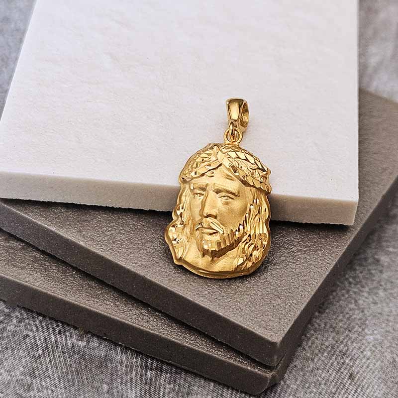 A gold Jesus Christ pendant with a textured crown of thorns, resting on a white marble block and dark gray stone blocks.