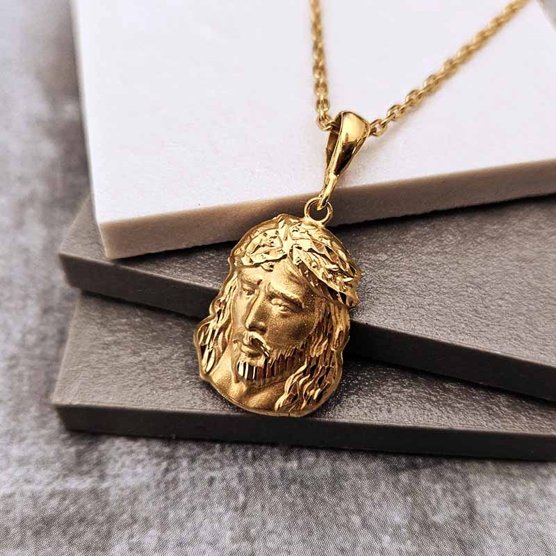 A gold Jesus Christ pendant necklace with a detailed face and crown of thorns, resting on stacked gray and white display blocks.