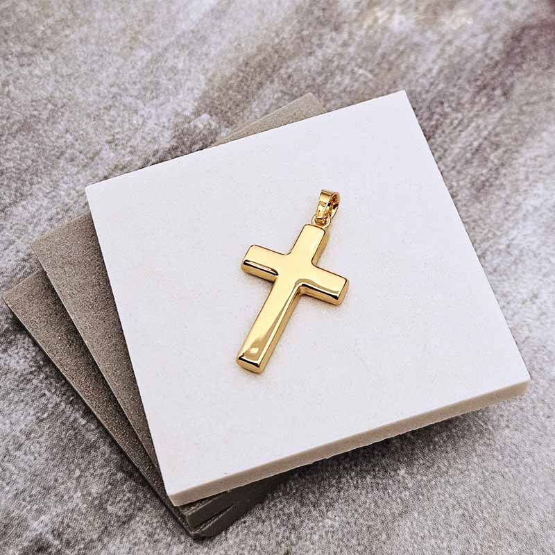 A polished gold cross pendant displayed on a white square card, resting on stacked gray cards against a textured gray background.