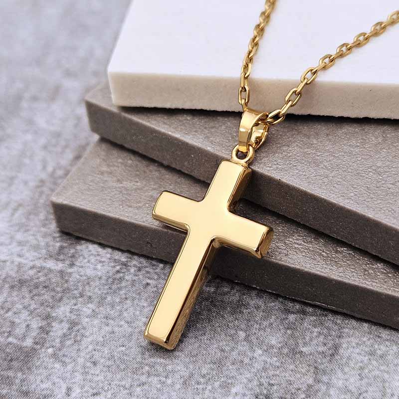 A polished gold cross pendant necklace on a matching gold chain, displayed on stacked gray and white blocks against a textured gray background.