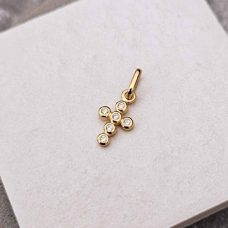 A small gold cross pendant charm with six clear round-cut gemstones, displayed on a textured white square card.