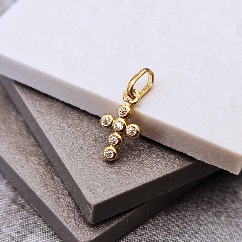 A gold cross pendant charm with small clear gemstones, displayed on stacked gray and white textured blocks.
