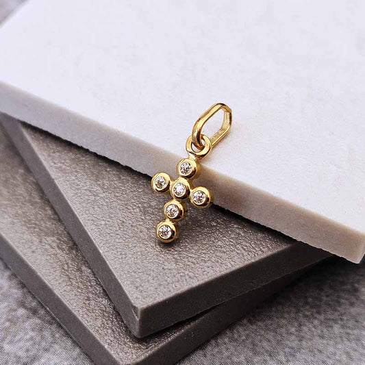 A gold cross pendant charm with small clear gemstones, displayed on stacked gray and white textured blocks.