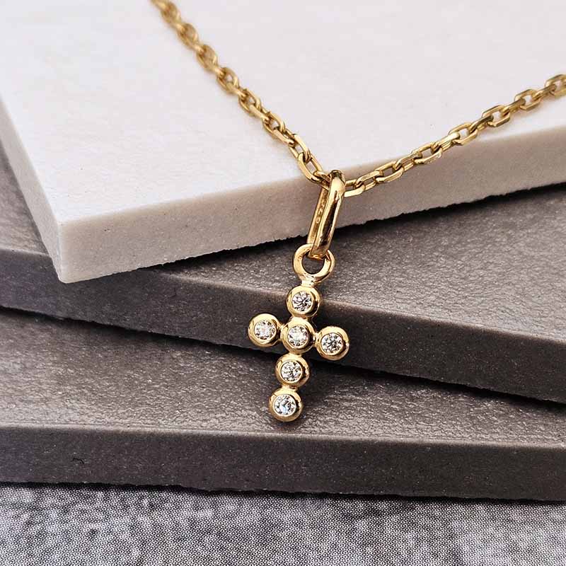 A gold cross pendant necklace with six clear round-cut stones, resting on a white marble block and dark gray textured stone surface.