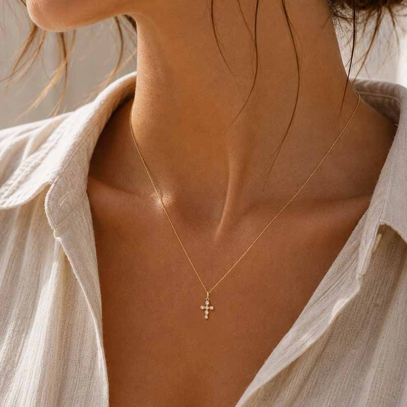 A close-up of a woman's neck and upper chest wearing a delicate gold cross pendant necklace with small pearl accents, paired with a light beige button-up shirt.