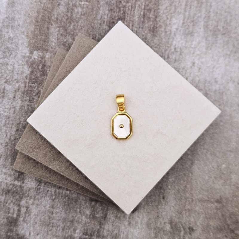A small gold pendant with a white inlay, displayed on a stack of neutral-colored square cards over a textured gray surface.