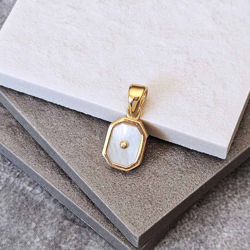 A gold pendant with an octagonal bezel containing a white mother-of-pearl inlay and a small central gold dot, displayed on stacked gray and white textured stone tiles.