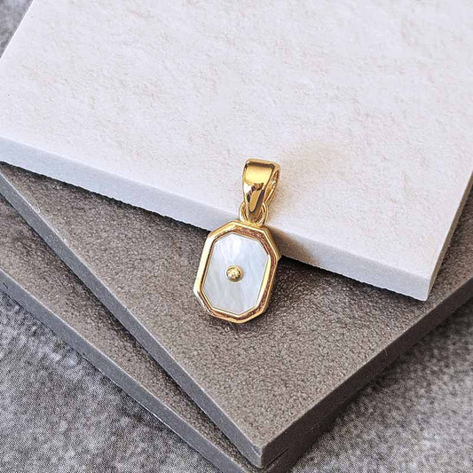 A gold pendant with an octagonal bezel containing a white mother-of-pearl inlay and a small central gold dot, displayed on stacked gray and white textured stone tiles.