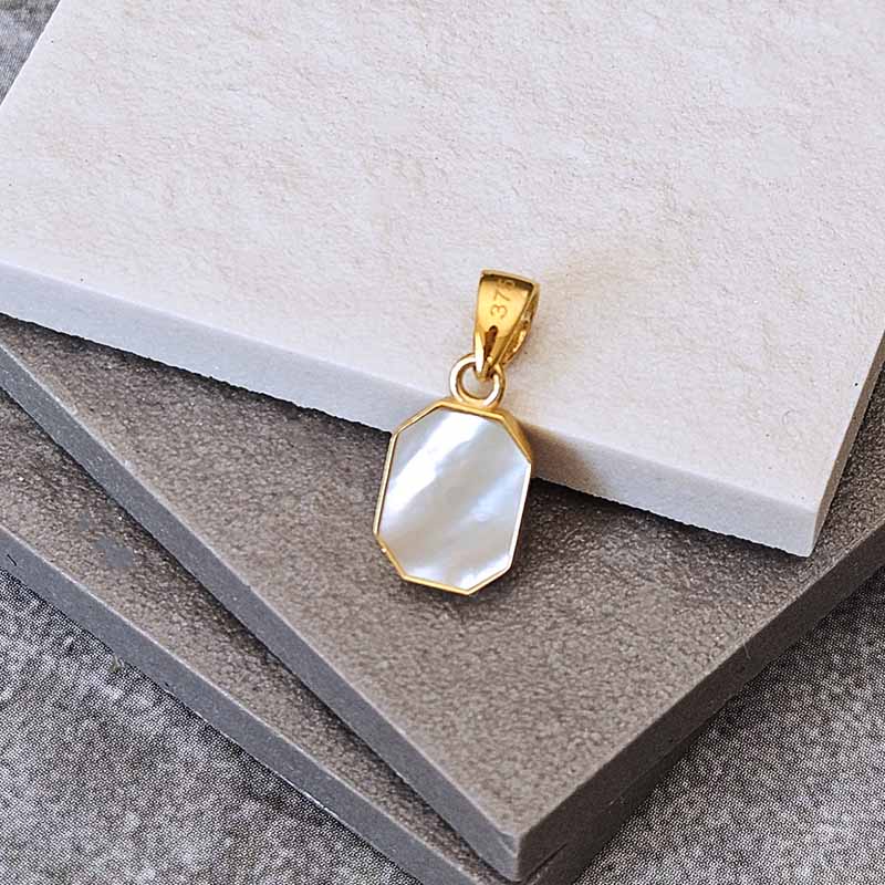 A gold pendant with an octagonal white mother-of-pearl inlay, displayed on stacked textured gray and off-white stone tiles.
