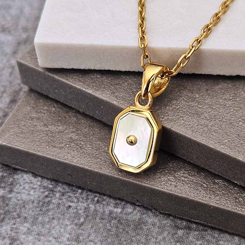 A gold chain necklace with an octagonal pendant featuring a white mother-of-pearl inlay and a small central gold dot, displayed on stacked gray and white blocks.
