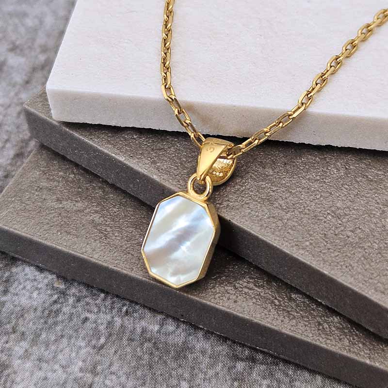 Gold chain necklace with a gold bezel-set octagonal mother-of-pearl pendant, displayed on stacked gray and white blocks.