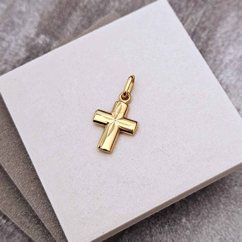 A polished gold cross pendant with a raised central star detail, displayed on a white square jewelry card over a gray textured surface.
