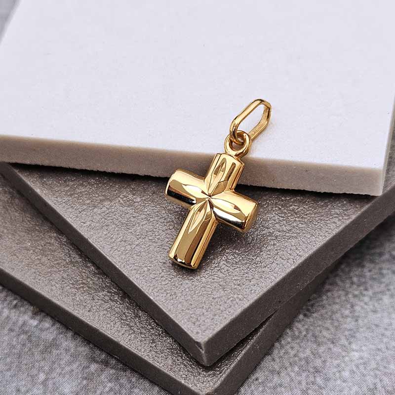 A polished gold cross pendant with a textured, ribbed surface and a hanging loop, displayed on a dark gray textured surface with a white card in the background.