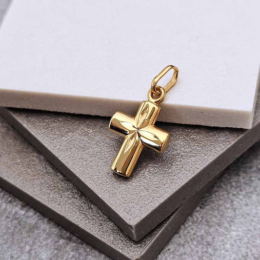 A polished gold cross pendant with a textured, ribbed surface and a hanging loop, displayed on a dark gray textured surface with a white card in the background.