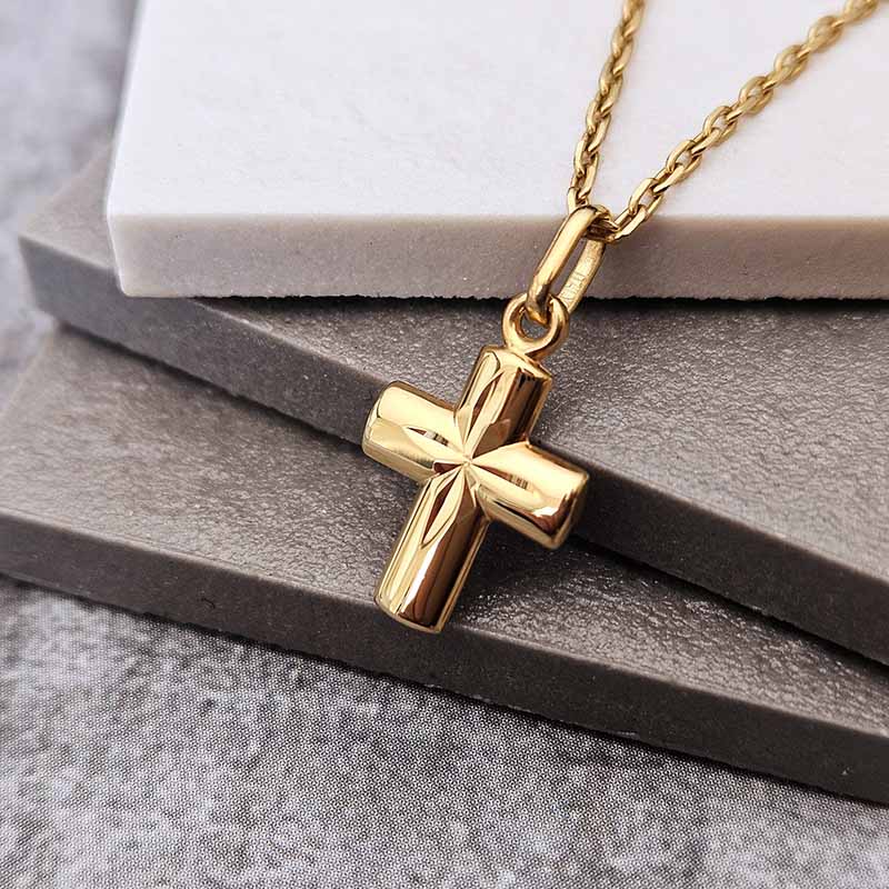 A polished gold cross pendant necklace on a gold chain, displayed on stacked gray and white stone blocks.