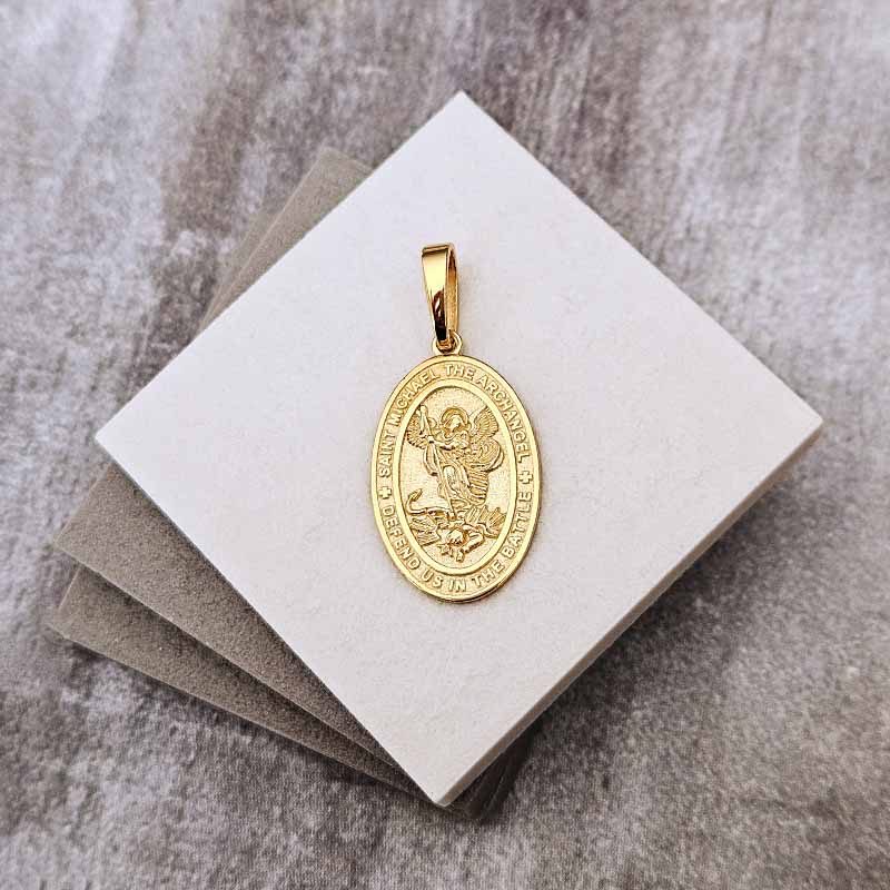 A gold oval pendant featuring a religious figure, displayed on a stack of white and gray square cards over a textured gray surface.